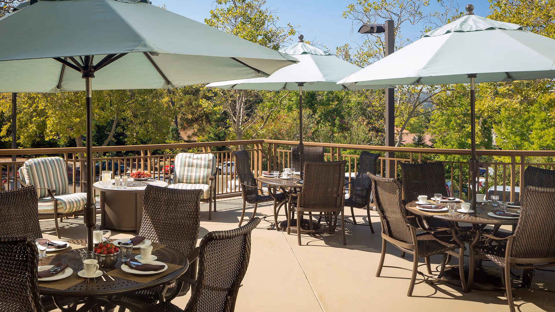 Outdoor patio area with several round tables and chairs under large white umbrellas. Tables are set with cups, glasses, and plates. The patio is surrounded by a railing and trees with green foliage are visible in the background under a clear blue sky.