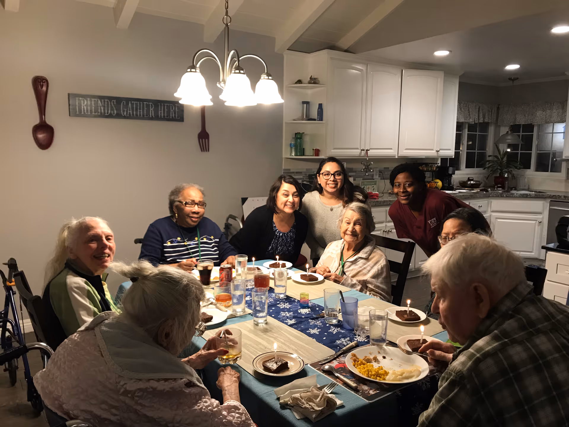 A group of elderly people and caregivers gathered around a dining table in a well-lit kitchen. The table is set with plates of food and desserts with lit candles. The wall has a sign that reads 'FRIENDS GATHER HERE' and large decorative utensils. The atmosphere is warm and social.
