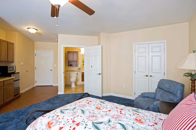 A bedroom with a floral patterned bedspread, a blue armchair, a wooden lamp with a beige lampshade, and a ceiling fan. The room has beige walls and blue carpet. There is an open door leading to a bathroom with a visible toilet and sink, and two closed white double doors on the right side.