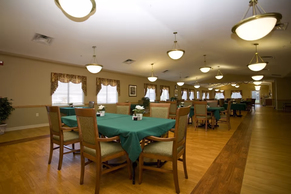 Spacious dining room with multiple tables covered in green tablecloths and wooden chairs under hanging pendant lights.