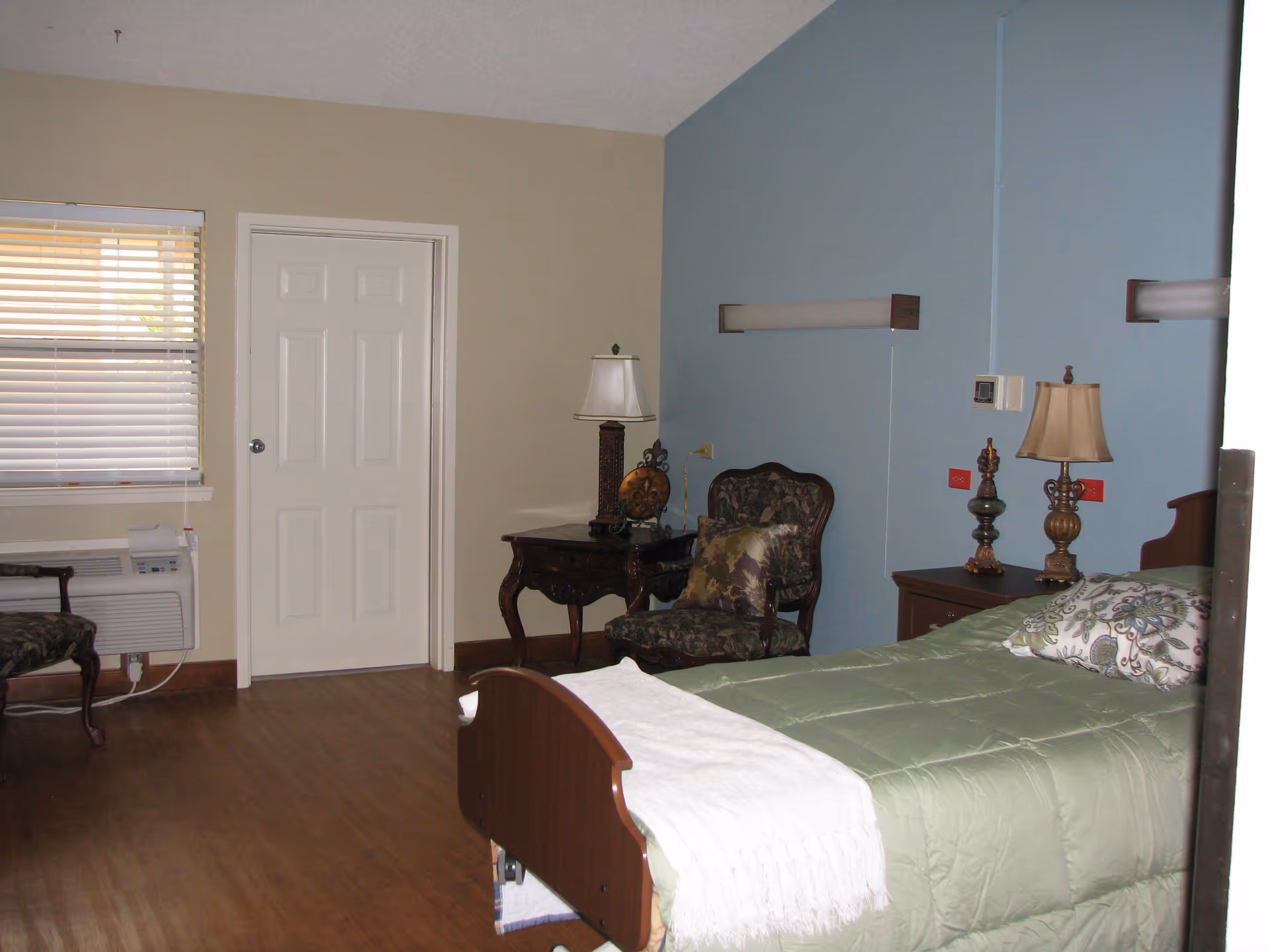 A bedroom in a senior living facility with a single bed covered in a green comforter and a white throw blanket. There are two decorative pillows on the bed. Next to the bed is a wooden nightstand with two ornate lamps and a small decorative item. An upholstered armchair with a floral pattern is positioned near a wooden side table with a lamp and a clock. The walls are painted beige and blue, and there is a window with blinds and an air conditioning unit below it. A closed white door is visible on the beige wall.
