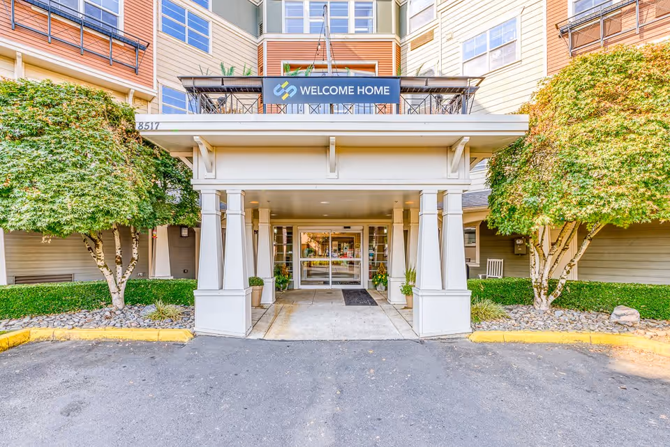 Entrance of a senior living facility with a covered driveway supported by white pillars, flanked by green trees and landscaping. Above the entrance is a sign that reads 'WELCOME HOME'. The building has multiple windows and a beige exterior.