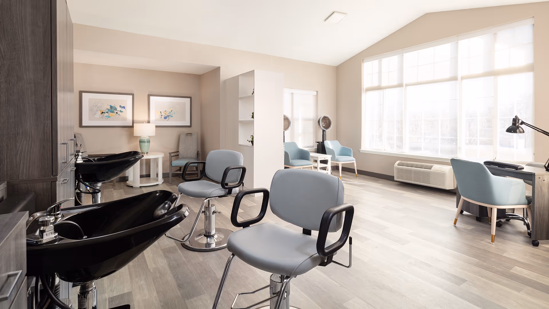 A bright and modern salon area with two black hair washing sinks and two gray salon chairs in the foreground. The room features large windows with white blinds letting in natural light, light wood flooring, and light-colored walls. In the background, there are additional seating areas with blue chairs, a small table with a lamp, and two framed abstract artworks on the wall.