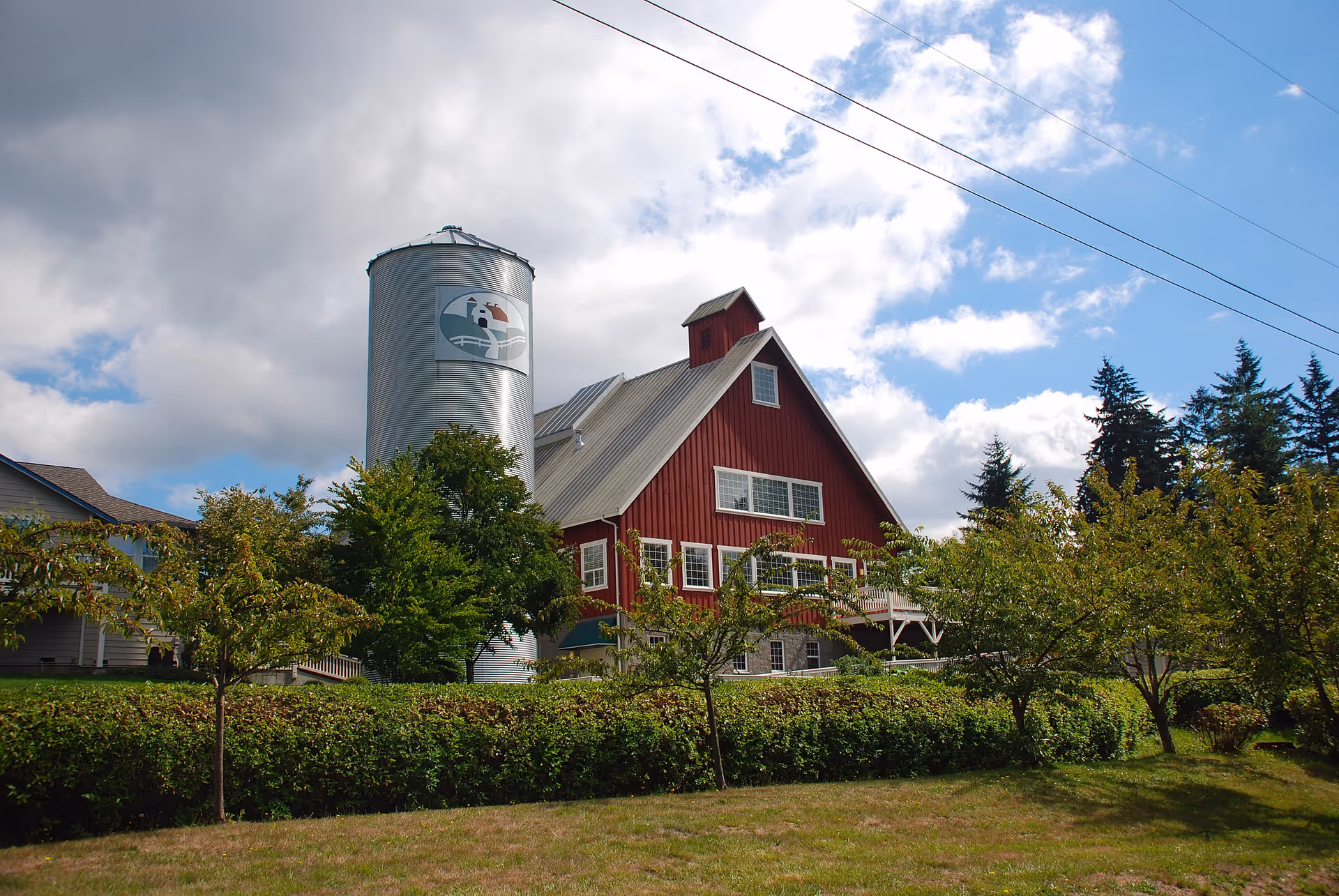 A large red barn-style building with a silver silo next to it, surrounded by trees and greenery under a partly cloudy blue sky.
