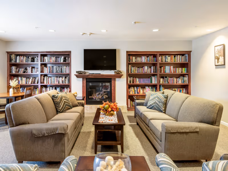 A cozy living room with two beige sofas facing each other across a wooden coffee table decorated with a fall-themed centerpiece. Behind the sofas are two large wooden bookshelves filled with books, a fireplace with a wooden mantel, and a flat-screen TV mounted above the fireplace. The room is well-lit with recessed ceiling lights and has a neutral color scheme.