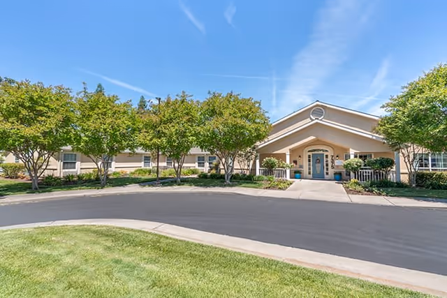 Front exterior view of Brookdale Sylvan Ranch facility with a paved driveway, green lawn, and several trees lining the front entrance under a clear blue sky.