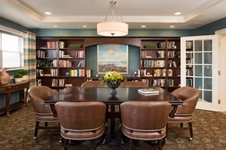 A cozy library or meeting room with a dark wooden table surrounded by six brown leather chairs. Behind the table is a large built-in bookshelf filled with books and decorative items, with a framed painting of a landscape in the center. The room has beige walls, a patterned carpet, a round ceiling light fixture, and a window with striped curtains on the left side. A small wooden side table with a plant and decorative vase is also visible.