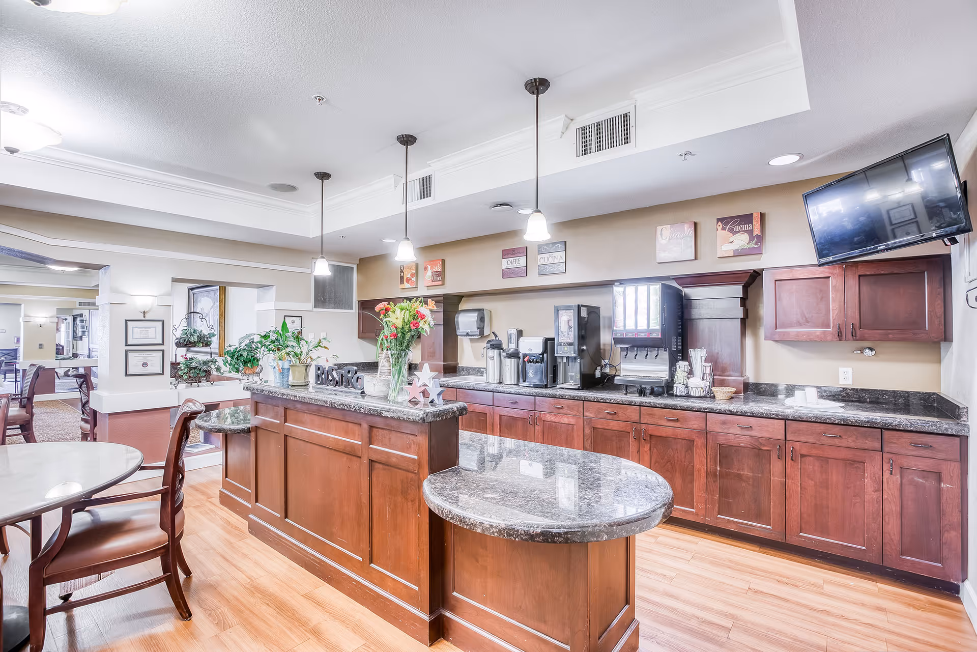 Interior view of a bright and clean assisted living facility common area featuring a coffee and beverage station with dark wood cabinets and granite countertops. There are three pendant lights hanging above the counter, a flat-screen TV mounted on the wall, and several decorative plants and flowers on the counter. In the background, there are tables and chairs for residents to sit and enjoy refreshments.
