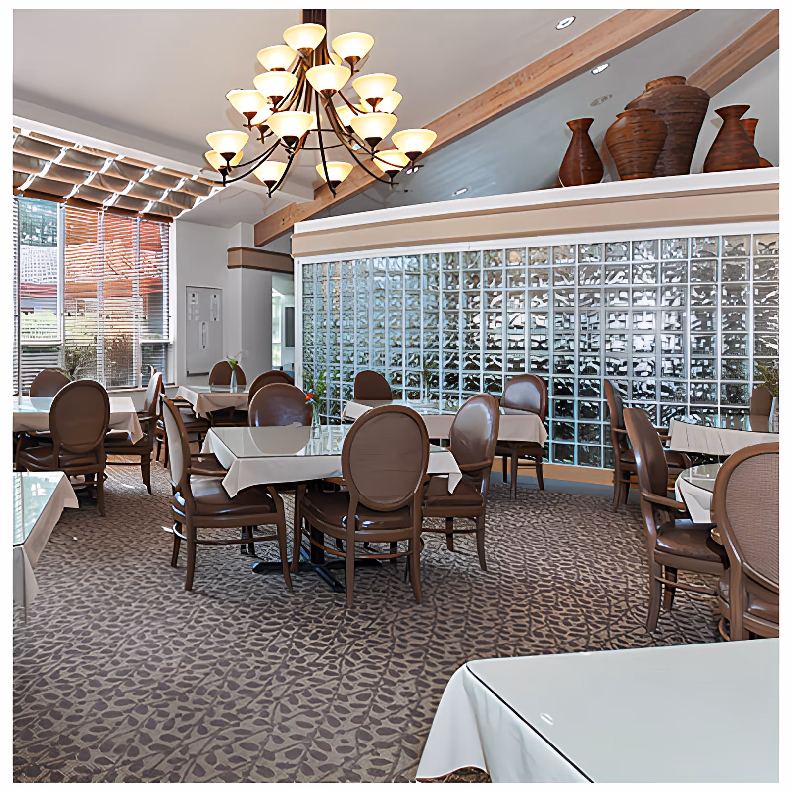 A dining room with multiple tables covered with white tablecloths and surrounded by brown chairs. The room features a large chandelier with multiple lights hanging from the ceiling, a patterned carpet, and a wall made of glass blocks. There are decorative vases on a ledge above the glass block wall and large windows with blinds letting in natural light.