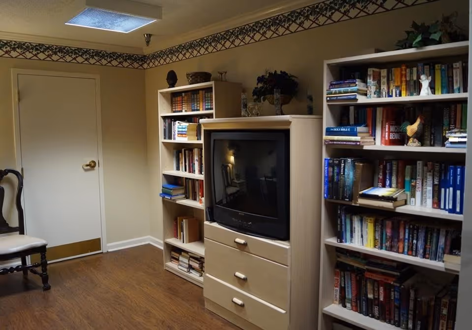 Interior room with wooden flooring, two bookshelves filled with books, a TV cabinet with an older style television, a wooden chair with a cushioned seat, and a closed door. The walls are beige with a decorative border near the ceiling.