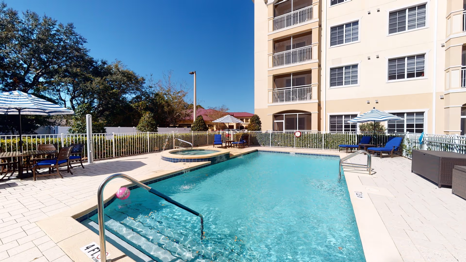 Outdoor swimming pool area at American House Bluewater Bay with clear blue water, a hot tub, lounge chairs with blue cushions, tables with blue and white striped umbrellas, and a multi-story building in the background under a clear blue sky.
