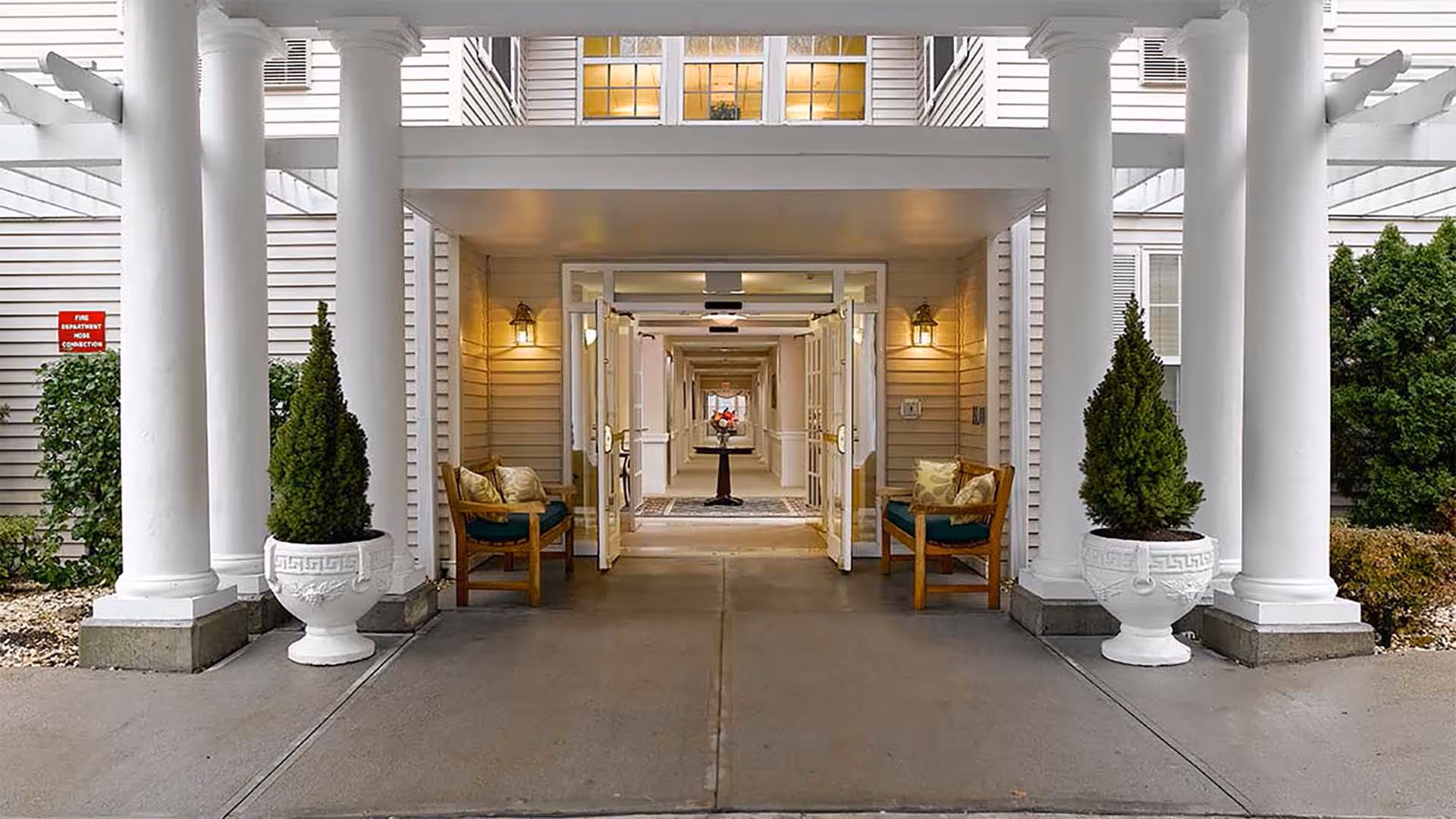 Entrance to a senior living facility with white columns and two potted plants on either side. There are two wooden chairs with cushions placed near the entrance. The double glass doors are open, revealing a long hallway with a table and flowers in the center.