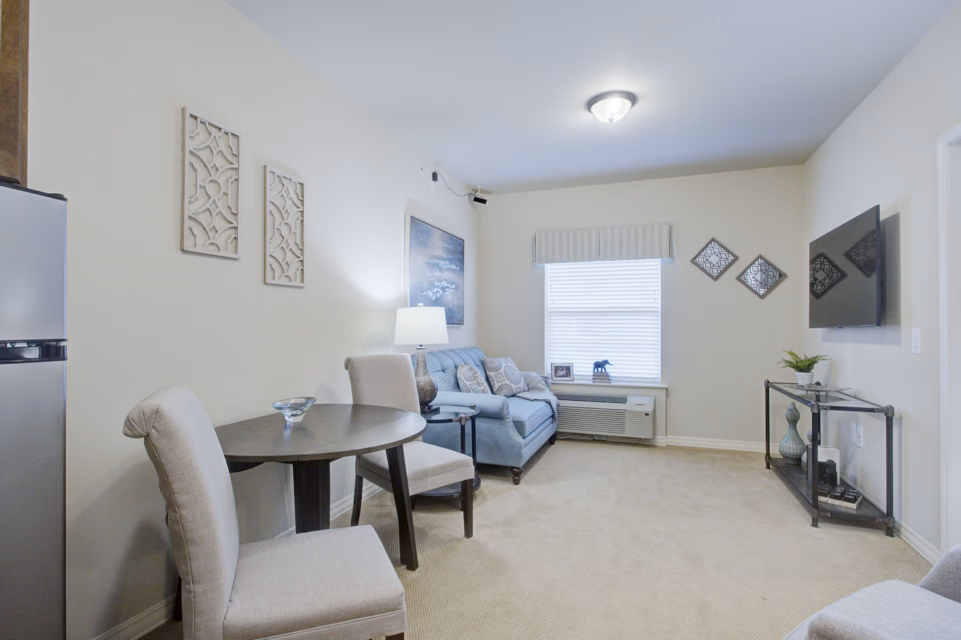 Cozy living room with a small round dining table and chairs, a blue sofa, wall-mounted TV and a window with blinds.