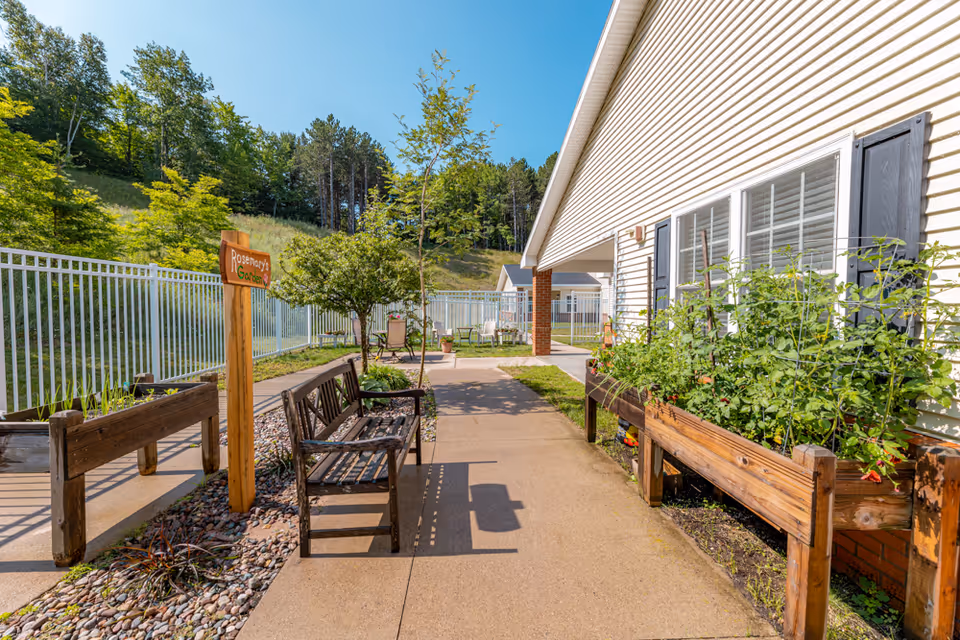 Outdoor garden area at Brookridge Heights featuring raised wooden garden beds with plants, a wooden bench, a small tree, and a sign that reads 'Rosemary's Garden'. The area is paved with a concrete walkway and enclosed by a white metal fence, with a building on the right side and trees in the background under a clear blue sky.