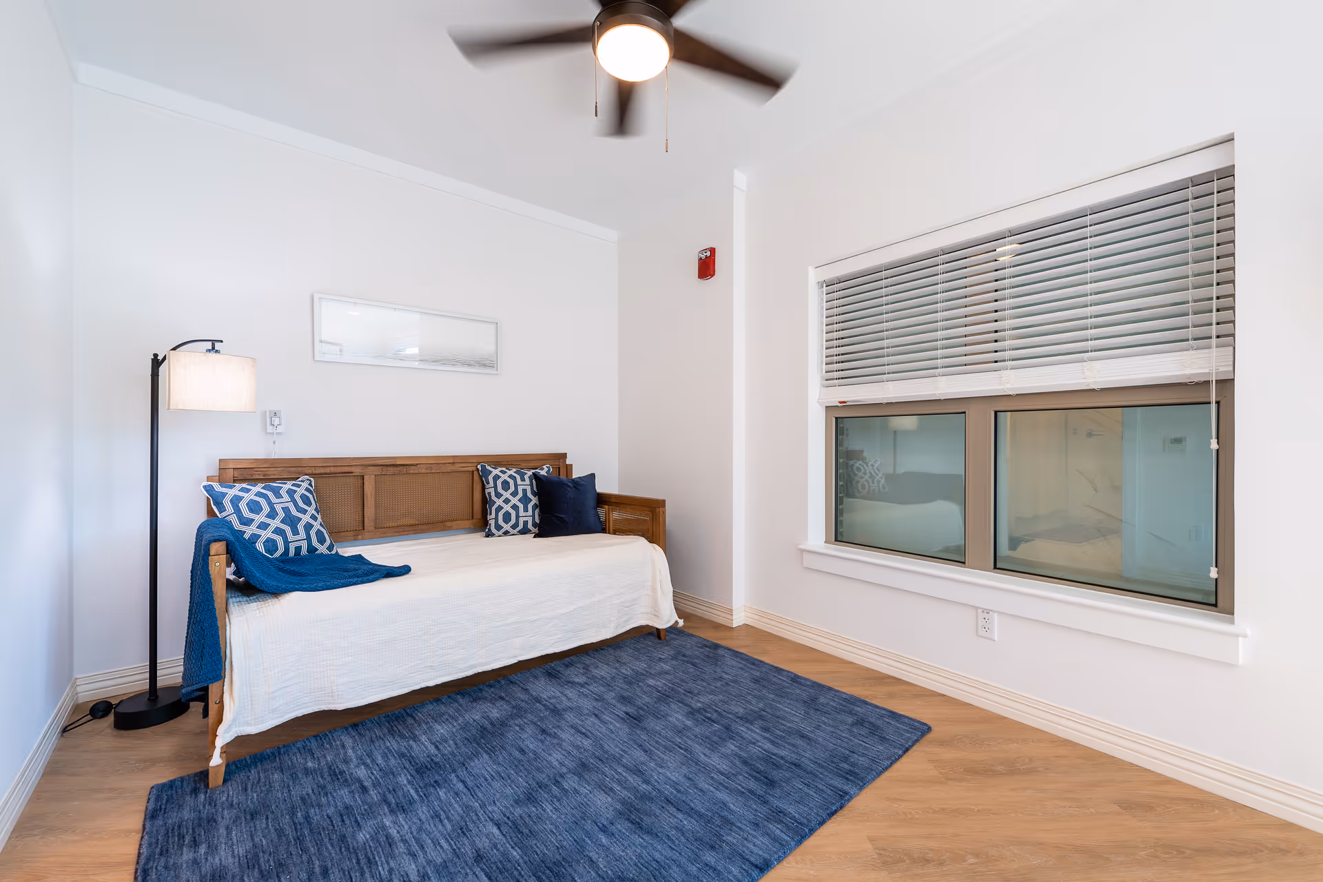 A bright room with a wooden daybed covered with a white blanket and decorated with blue and patterned pillows. A blue rug lies on the wooden floor in front of the daybed. A tall floor lamp stands to the left of the daybed, and a ceiling fan with a light is mounted above. There is a window with closed blinds on the right wall.