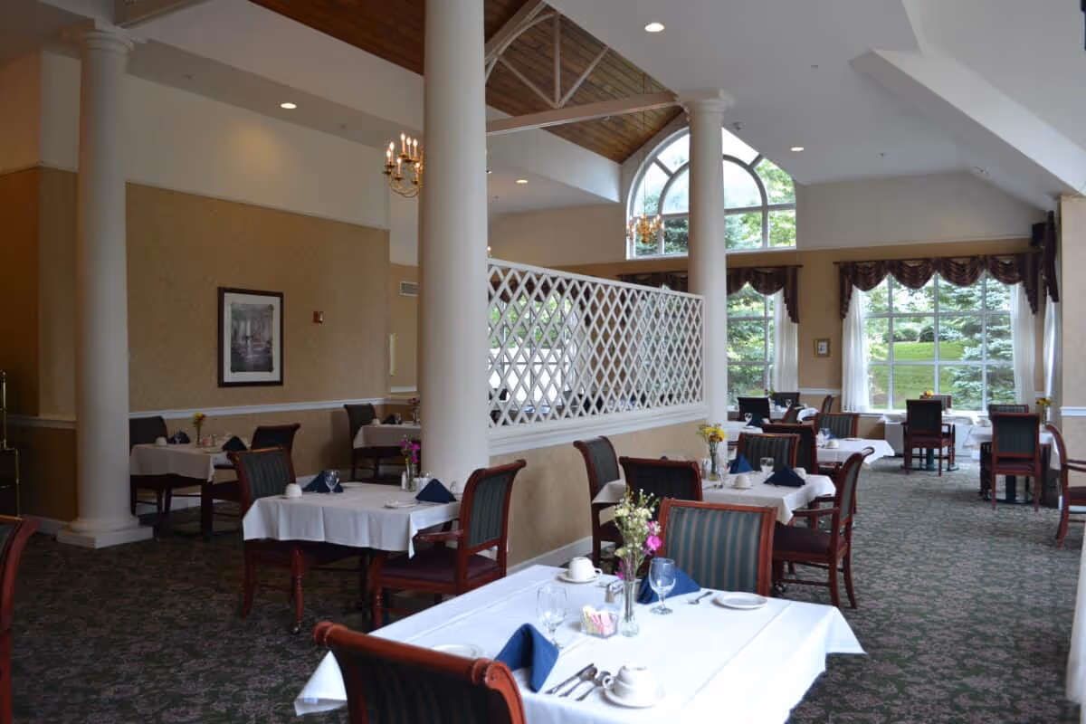 A bright and elegant dining room with multiple tables covered in white tablecloths, set with cups, glasses, and napkins. The room features large windows with views of greenery outside, tall white columns, a chandelier, and a decorative lattice partition.