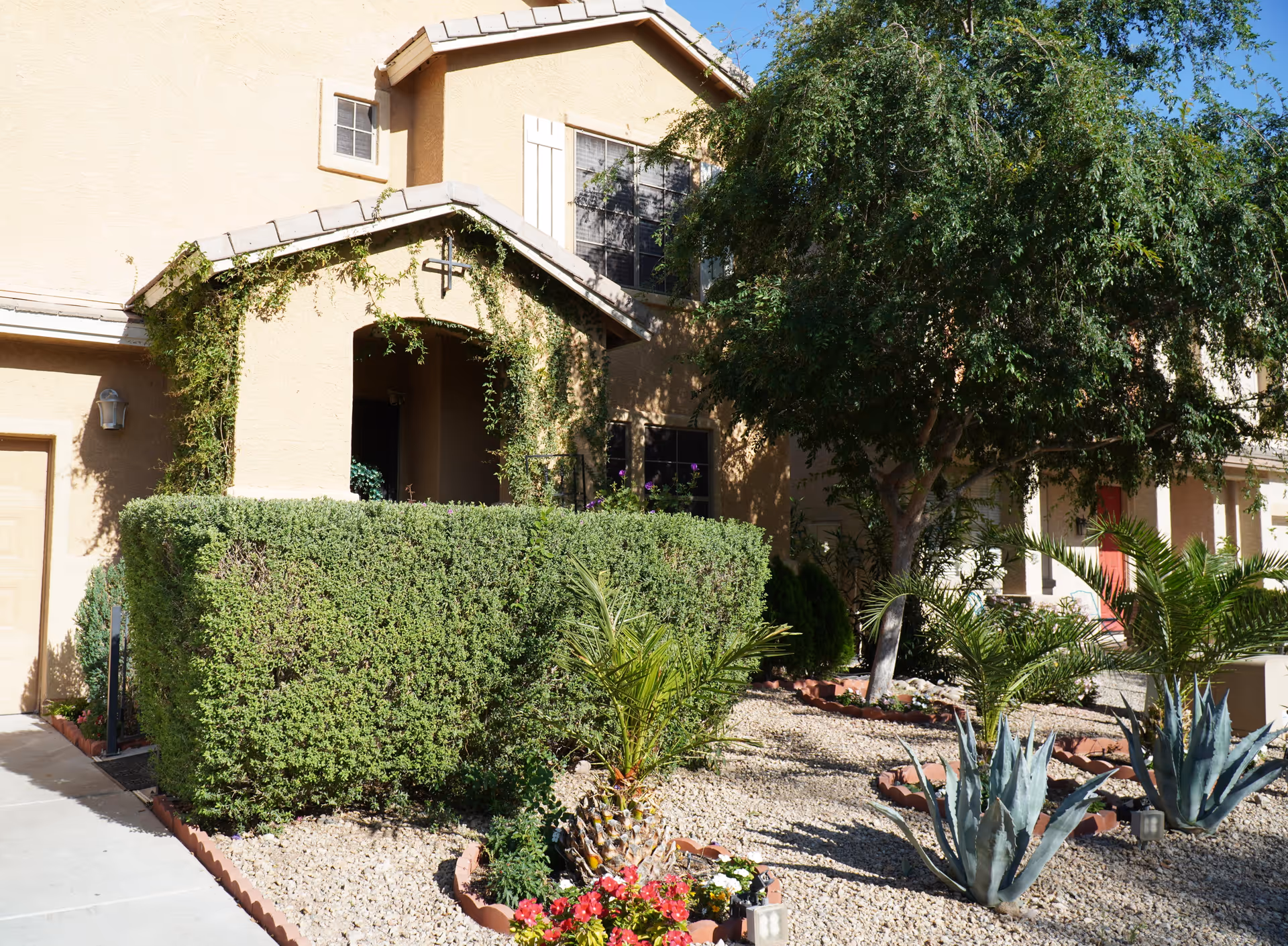 Exterior view of a beige stucco assisted living home with a small covered porch, surrounded by neatly trimmed bushes, various desert plants, and a tree providing shade.
