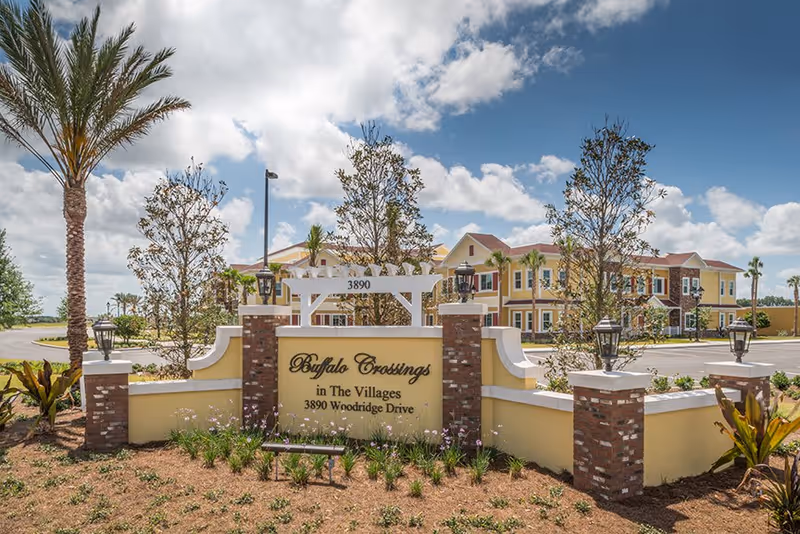 Entrance sign for Buffalo Crossings in The Villages at 3890 Woodridge Drive, with a landscaped area featuring palm trees and plants, and a large two-story yellow building in the background under a partly cloudy sky.