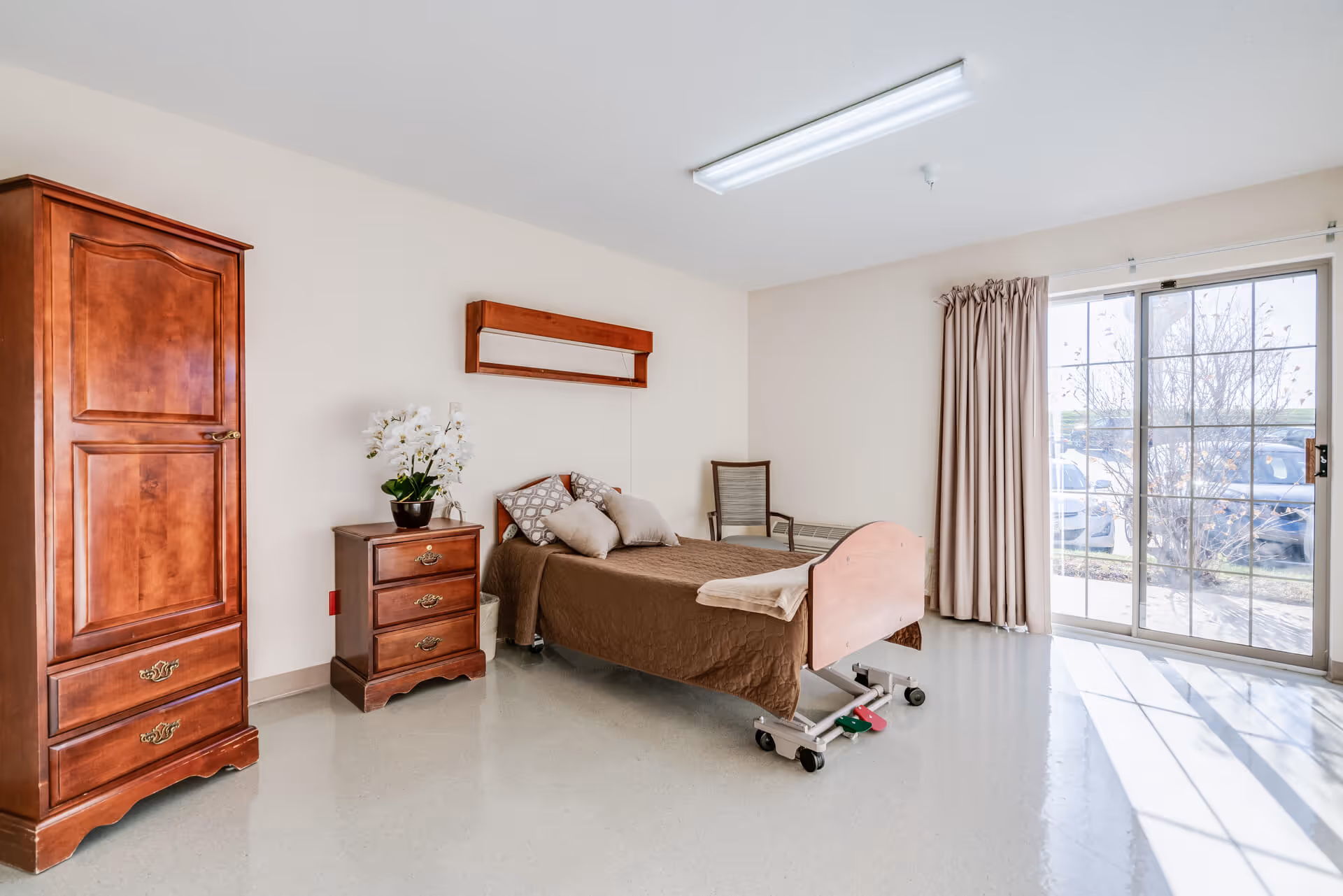 A senior living facility bedroom with a single bed covered in a brown quilt and several pillows. Next to the bed is a wooden nightstand with a white orchid plant on top. There is a wooden wardrobe to the left and a chair in the corner. The room has a large sliding glass door with beige curtains, letting in natural light.
