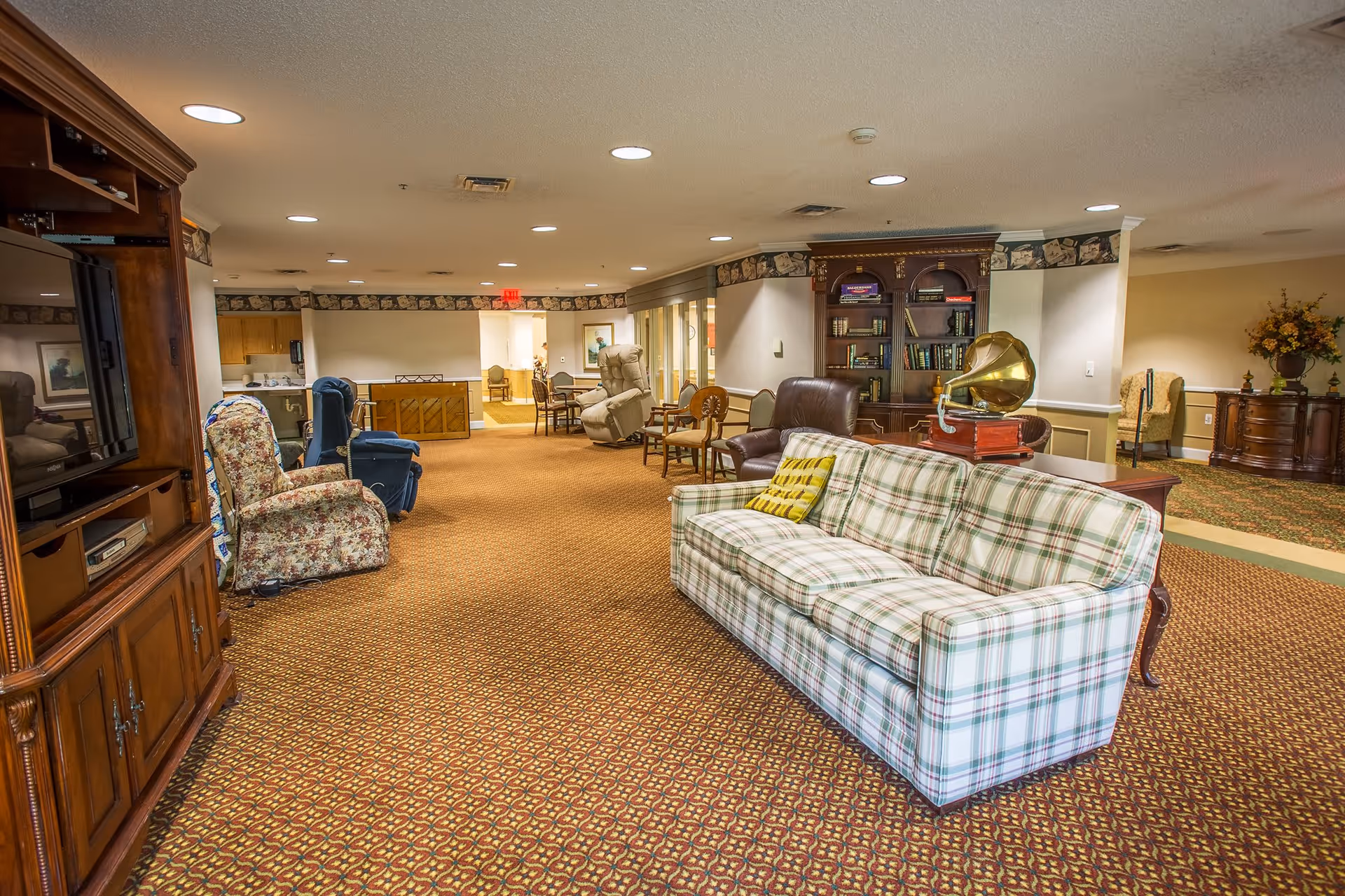 A spacious senior living facility common area with patterned carpet, a plaid sofa with a yellow pillow, various armchairs, a wooden entertainment center with a TV, a bookshelf, and a vintage gramophone. The room is well-lit with recessed ceiling lights and decorated with floral arrangements and framed artwork.