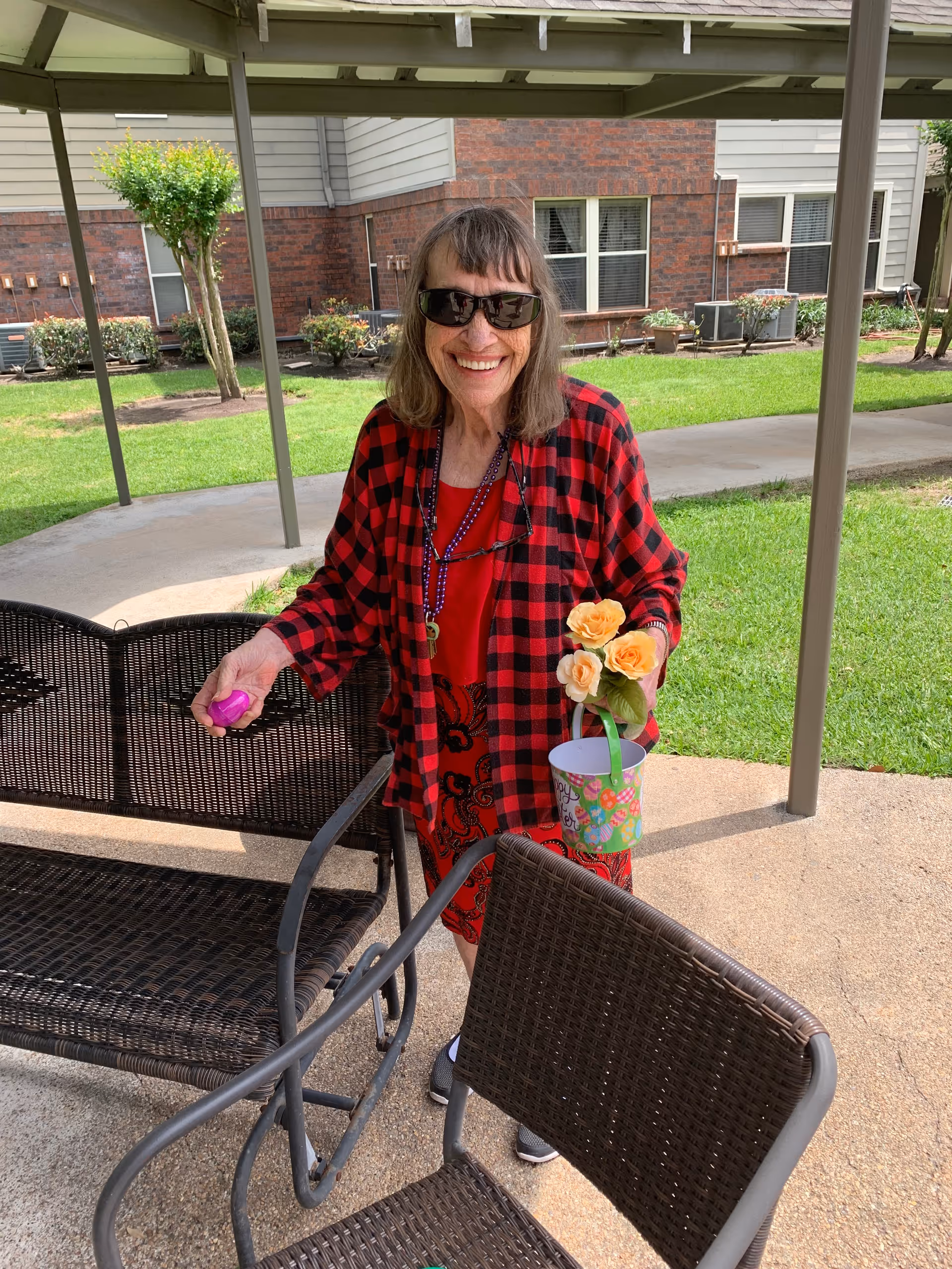 An elderly woman wearing sunglasses and a red and black checkered shirt stands outdoors under a covered patio. She is smiling and holding a small colorful bucket with yellow flowers in one hand and a pink object in the other. Behind her is a grassy area with bushes and a building with brick and siding.