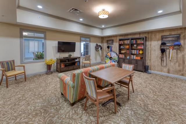 A cozy common area with patterned carpet, a striped sofa, and a wooden table with four chairs. The room features two windows, a wall-mounted TV above a wooden media console, a bookshelf filled with books and decorative items, and a colorful prize wheel. Several hats and accessories hang on the wall, and the ceiling has recessed lighting and a central light fixture.