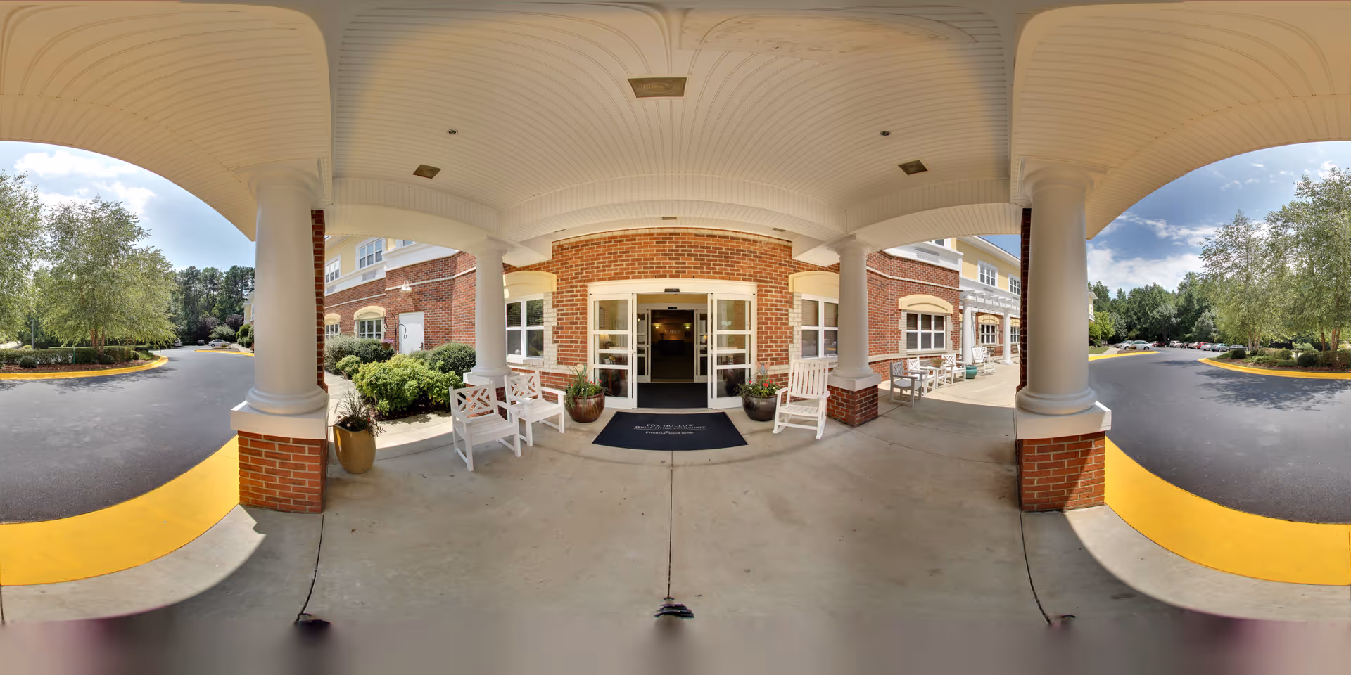 Covered entrance area of Fox Hollow Senior Living Community with white columns and brick walls. There are white chairs and potted plants on the concrete porch. The entrance has glass doors leading inside. Surrounding the entrance are landscaped bushes and trees, with a paved driveway curving around the building under a partly cloudy sky.