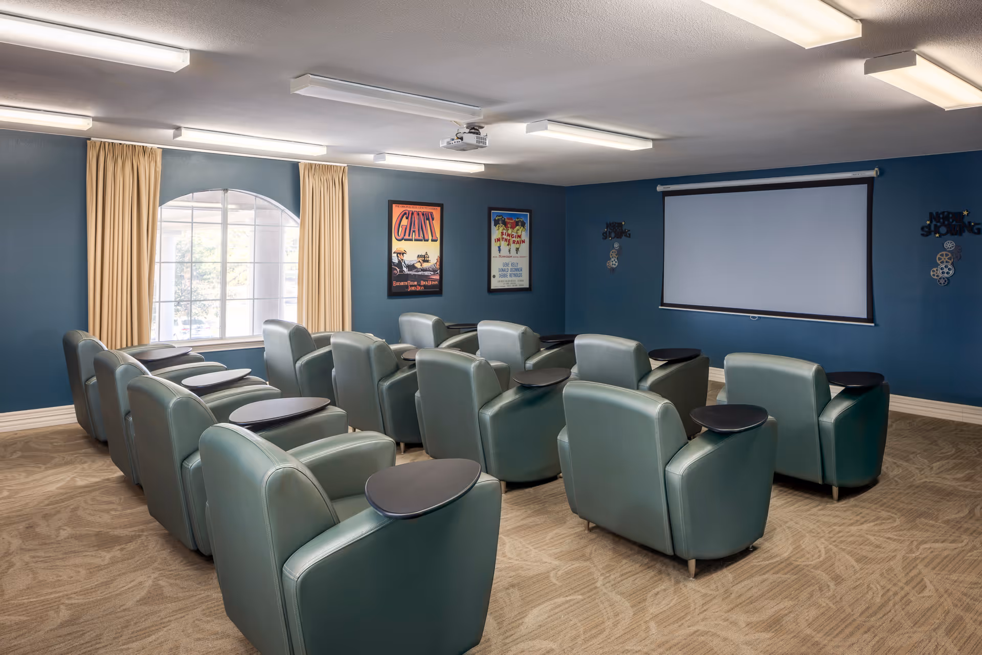 A small theater room with two rows of green leather recliner chairs, each equipped with a small black tray table. The room has blue walls, a large window with beige curtains, two framed movie posters, and a white projector screen at the front.
