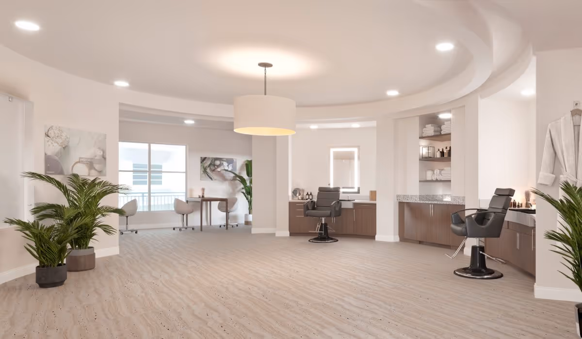 A spacious, modern salon area in a senior living facility with two black salon chairs in front of mirrors and wooden cabinets. The room features light-colored walls and flooring, potted plants, a round ceiling light fixture, and a small table with chairs near a window.