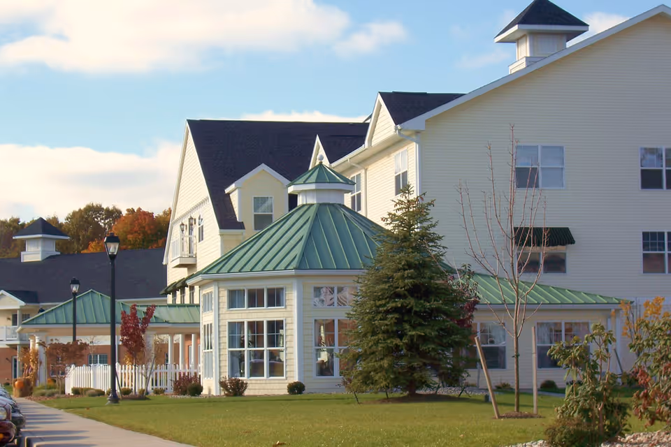 Exterior view of a senior living facility named Legacy at Clover Blossom, featuring a large light yellow building with green metal roofs, multiple windows, and a well-maintained lawn with trees and shrubs under a partly cloudy sky.
