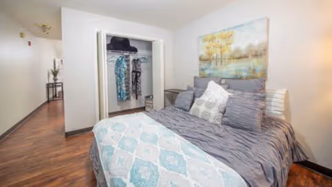 A cozy bedroom in a retirement facility featuring a neatly made bed with multiple pillows and a patterned blanket. There is a large painting of a serene landscape hanging above the bed. An open closet with hanging clothes is visible, and a hallway extends from the room with a small table and decorative items along the wall.
