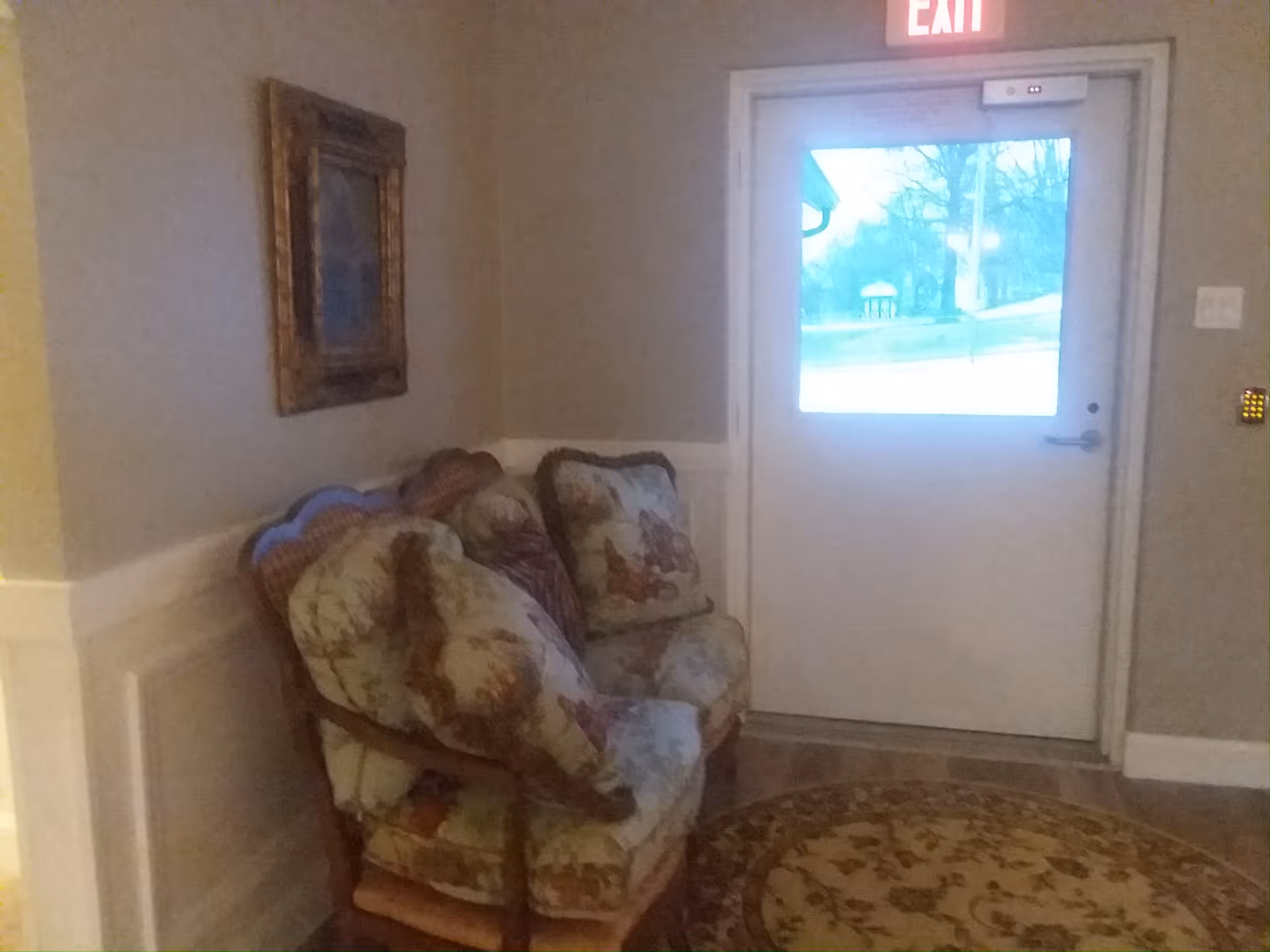 Interior entry area with floral-upholstered chairs, framed wall art, round rug and an exterior door marked EXIT.