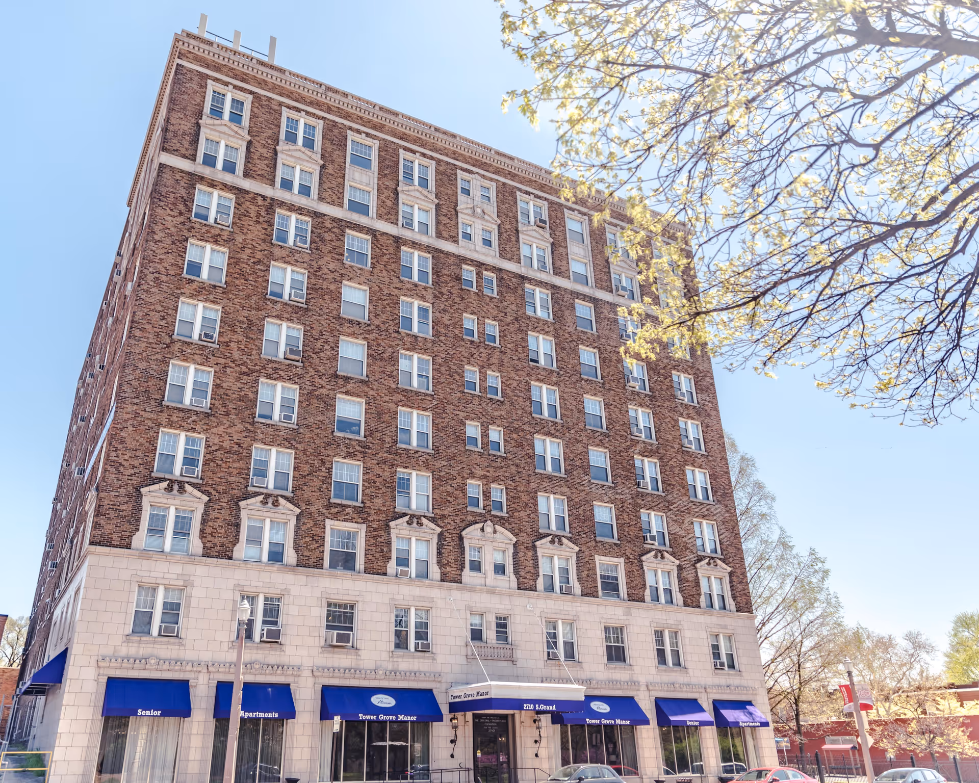 Exterior view of a multi-story brick building named Tower Grove Manor with blue awnings over the ground floor windows and entrance. Trees with budding leaves are visible on the right side under a clear blue sky.