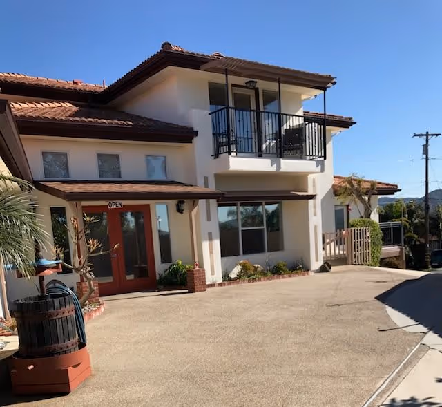 Exterior view of a two-story building with white walls and a red-tiled roof under a clear blue sky. The building has a balcony with black railings on the upper floor and large windows on the ground floor. There is a concrete driveway in front, a red double door entrance with an 'OPEN' sign above it, and some plants near the entrance.