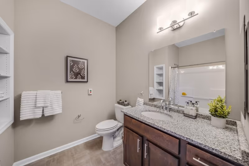 A bright, modern bathroom with a granite vanity and sink, large mirror, toilet, towel rack, and a bathtub with shower curtain.