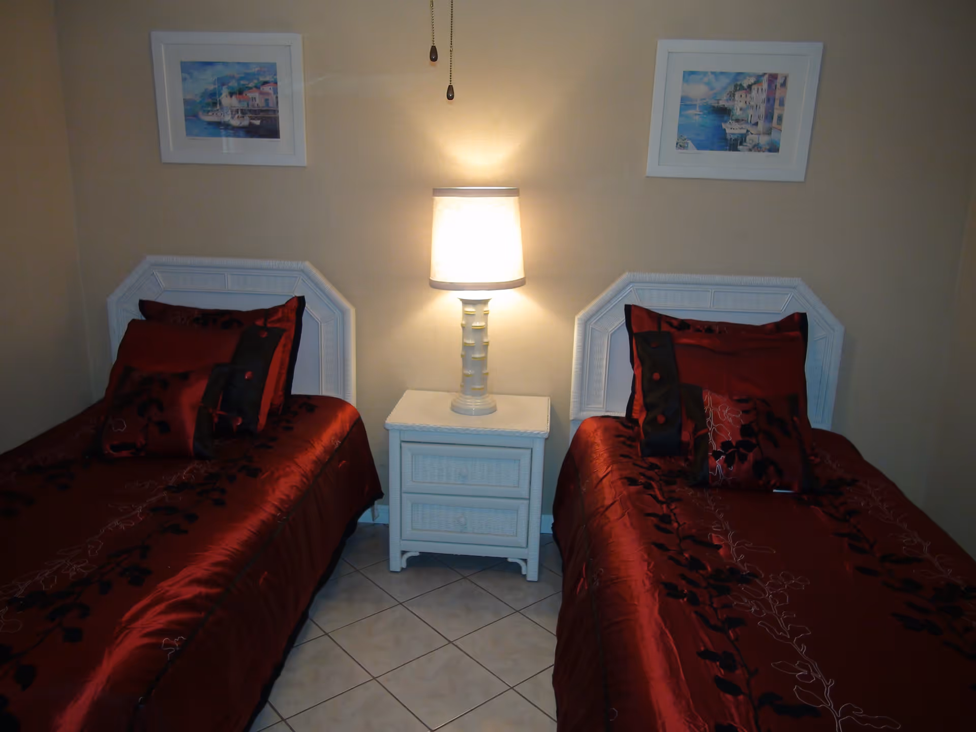 A bedroom with two twin beds featuring white headboards and red bedspreads with black floral patterns. A white nightstand with two drawers is placed between the beds, topped with a lit table lamp. Two framed paintings hang on the beige wall above each bed.