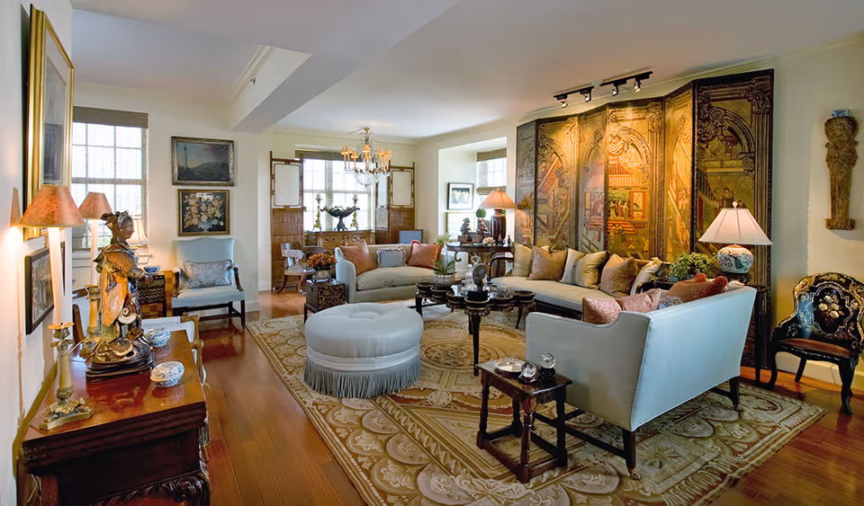 A spacious and elegantly decorated living room with a large area rug, multiple sofas and chairs, a decorative folding screen with intricate artwork, various lamps, and framed paintings on the walls. The room has wooden flooring and large windows allowing natural light to enter.