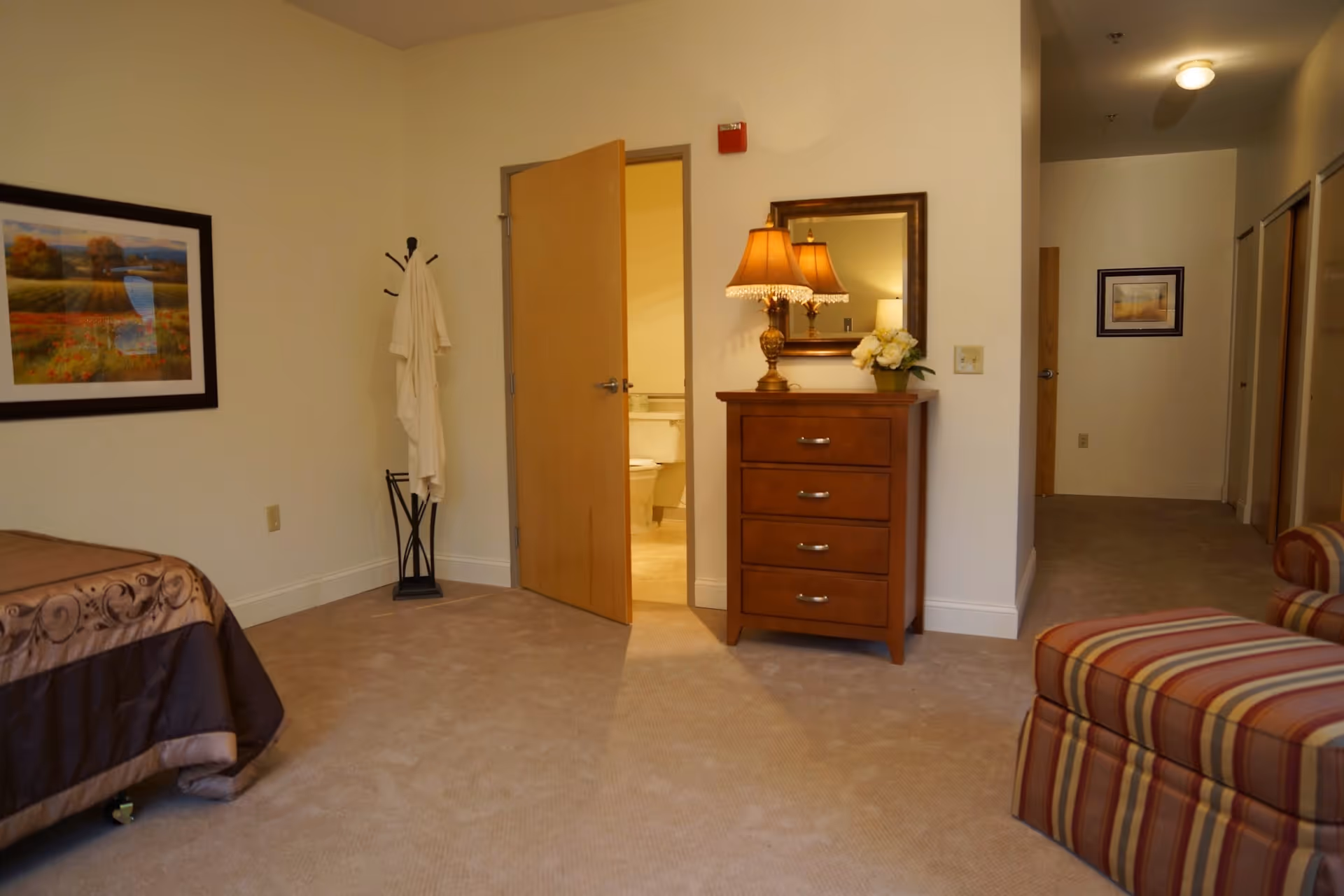 A cozy senior living bedroom with beige carpet, a bed partially visible on the left, a wooden dresser with a lamp and flowers on top, a mirror above the dresser, an open door leading to a bathroom, a coat rack with a white robe, and a striped armchair on the right side.