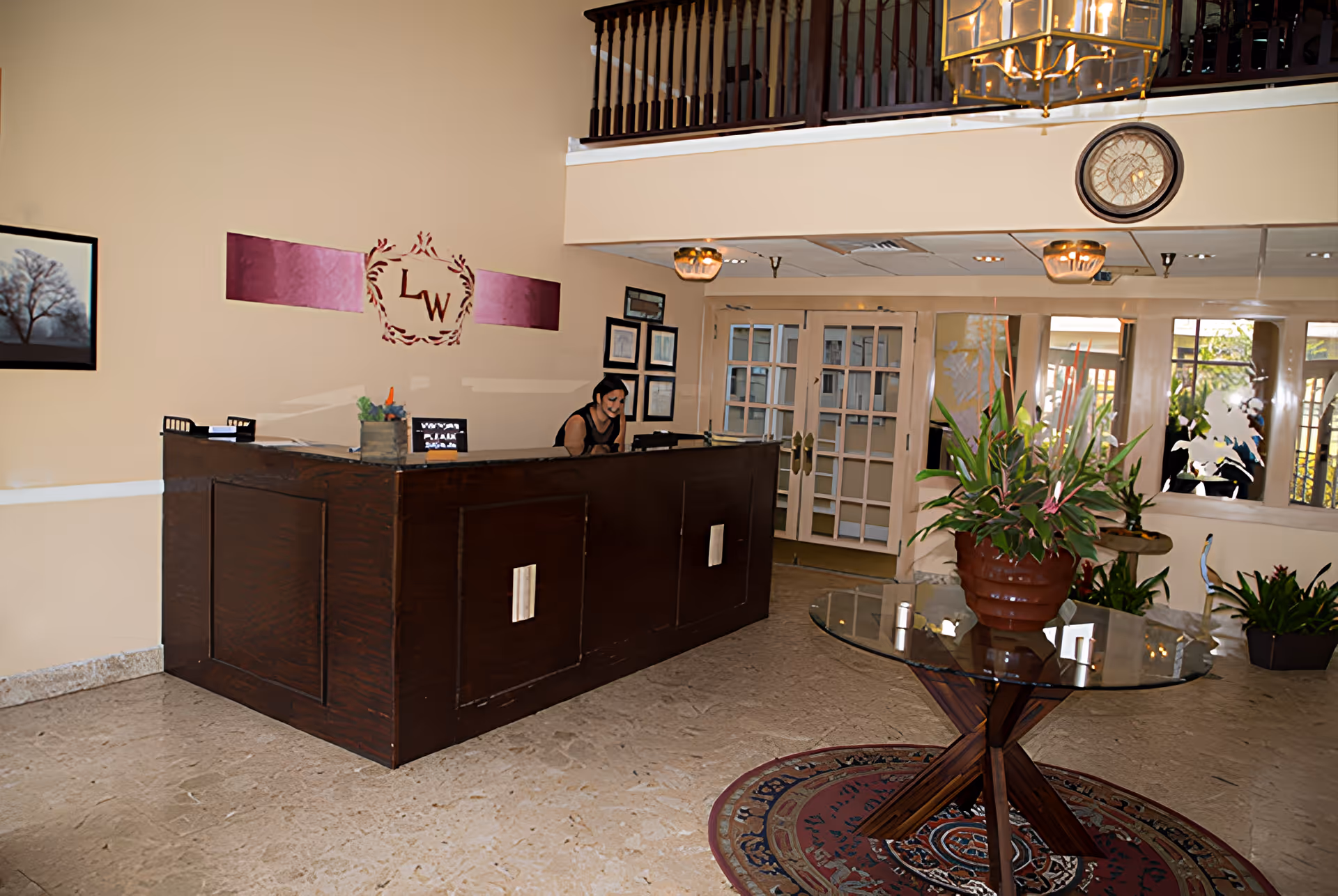 Reception area with a dark wooden front desk where a person is seated. Behind the desk is a beige wall with a decorative emblem featuring the letters 'LW'. To the right, there is a glass table with a large potted plant on a patterned rug. The area has beige tiled flooring, framed artwork on the walls, and glass double doors in the background. A balcony railing and a large hanging light fixture are visible above.