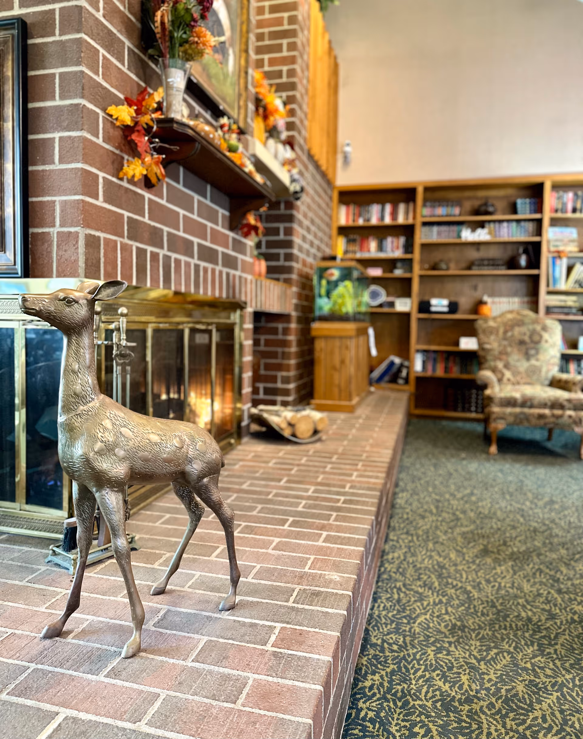 A cozy living room area with a brick fireplace decorated with autumn leaves and a mirror above the mantel. In front of the fireplace is a brass deer statue. To the right, there is a wooden bookshelf filled with books and an aquarium on a wooden stand. A patterned armchair is positioned near the bookshelf on a carpeted floor.
