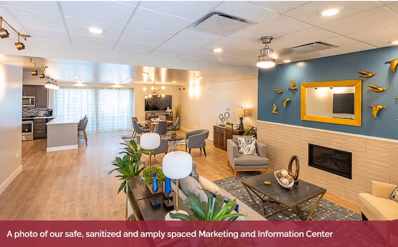 A spacious and well-lit common area in a senior living facility featuring a living room with comfortable chairs, a coffee table, decorative plants, a fireplace with a gold-framed mirror and bird wall art above it, and a dining area with a table and chairs near a kitchen with modern appliances and a breakfast bar.