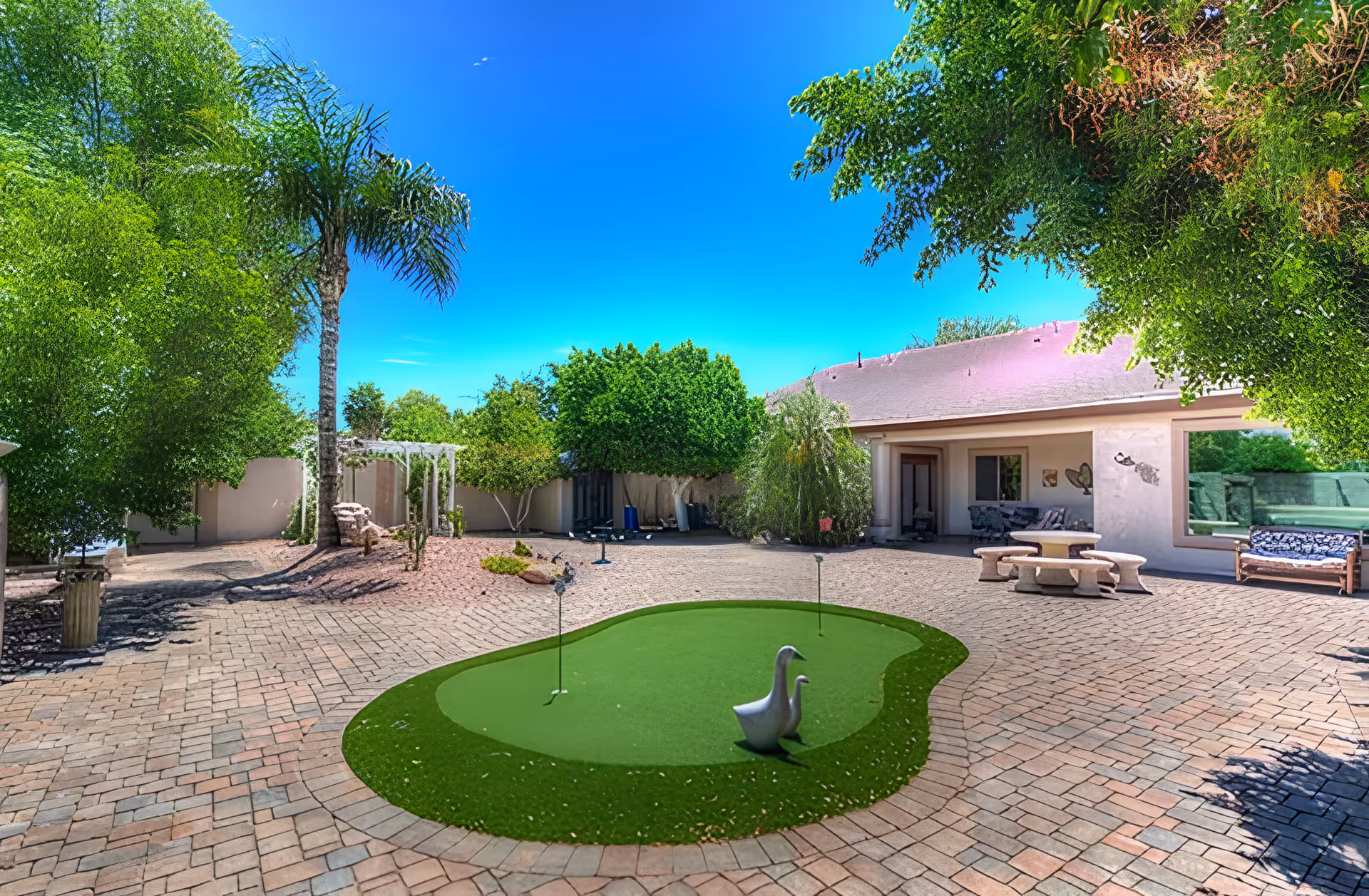 Outdoor courtyard area with a small putting green featuring two golf holes and two duck statues. The courtyard is paved with brick and surrounded by lush green trees and shrubs. There is a covered patio area with seating and a stone picnic table with benches. The sky is clear and blue.