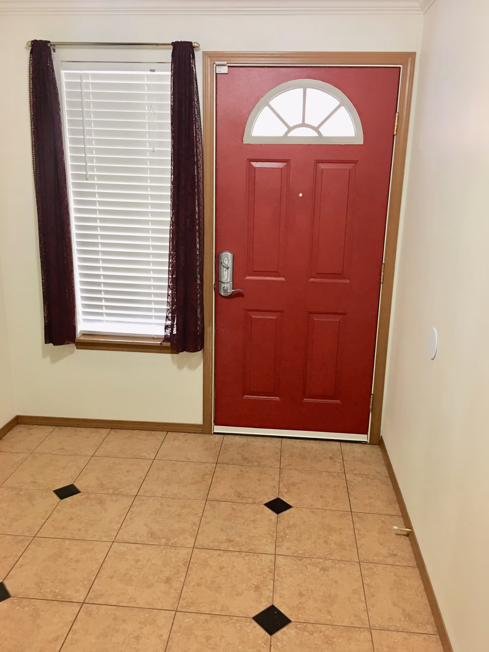 Interior view of an entryway with a red front door featuring a semi-circular window at the top. To the left of the door is a window with white blinds and dark red curtains. The floor is tiled with beige tiles accented by small black diamond-shaped tiles.