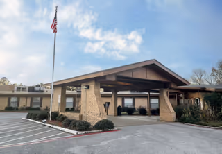 Front entrance of a single-story senior living facility with a covered porte-cochere, American flag, and parking area.
