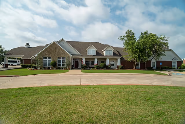 Front exterior of a one-story senior living facility with a covered entrance, driveway, and lawn.