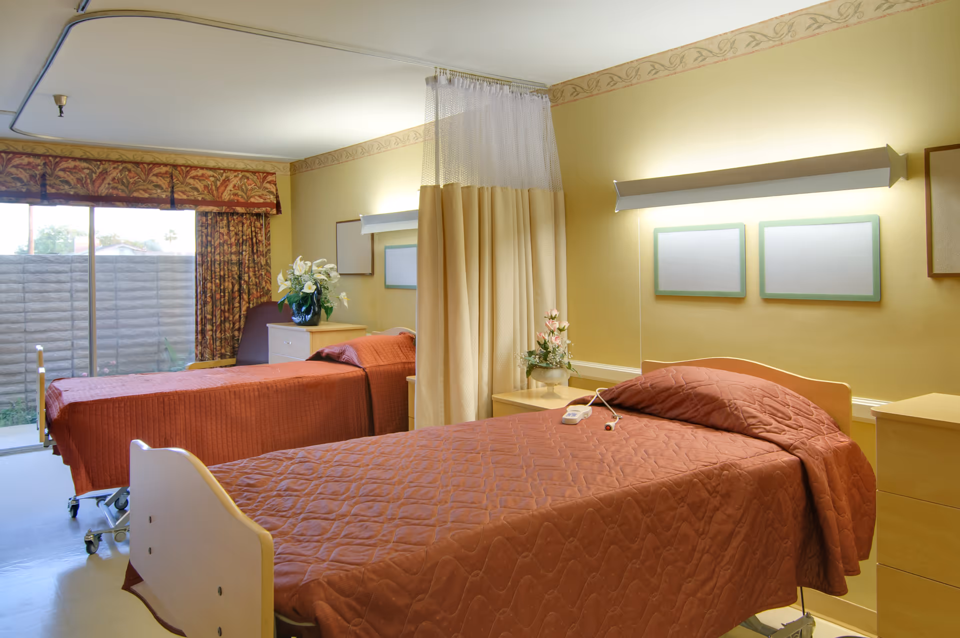 A hospital room with two single beds covered in brown quilts, separated by a beige privacy curtain. Each bed has a bedside table with flowers and a call button. The room has yellow walls, framed pictures, and a large window with patterned curtains letting in natural light.