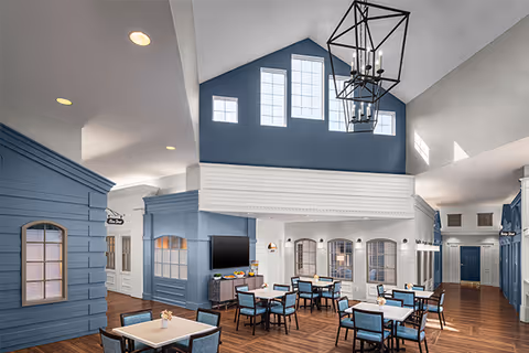 Bright high‑ceilinged communal dining area with multiple tables and chairs, blue and white interior, large windows, a TV and a central chandelier.