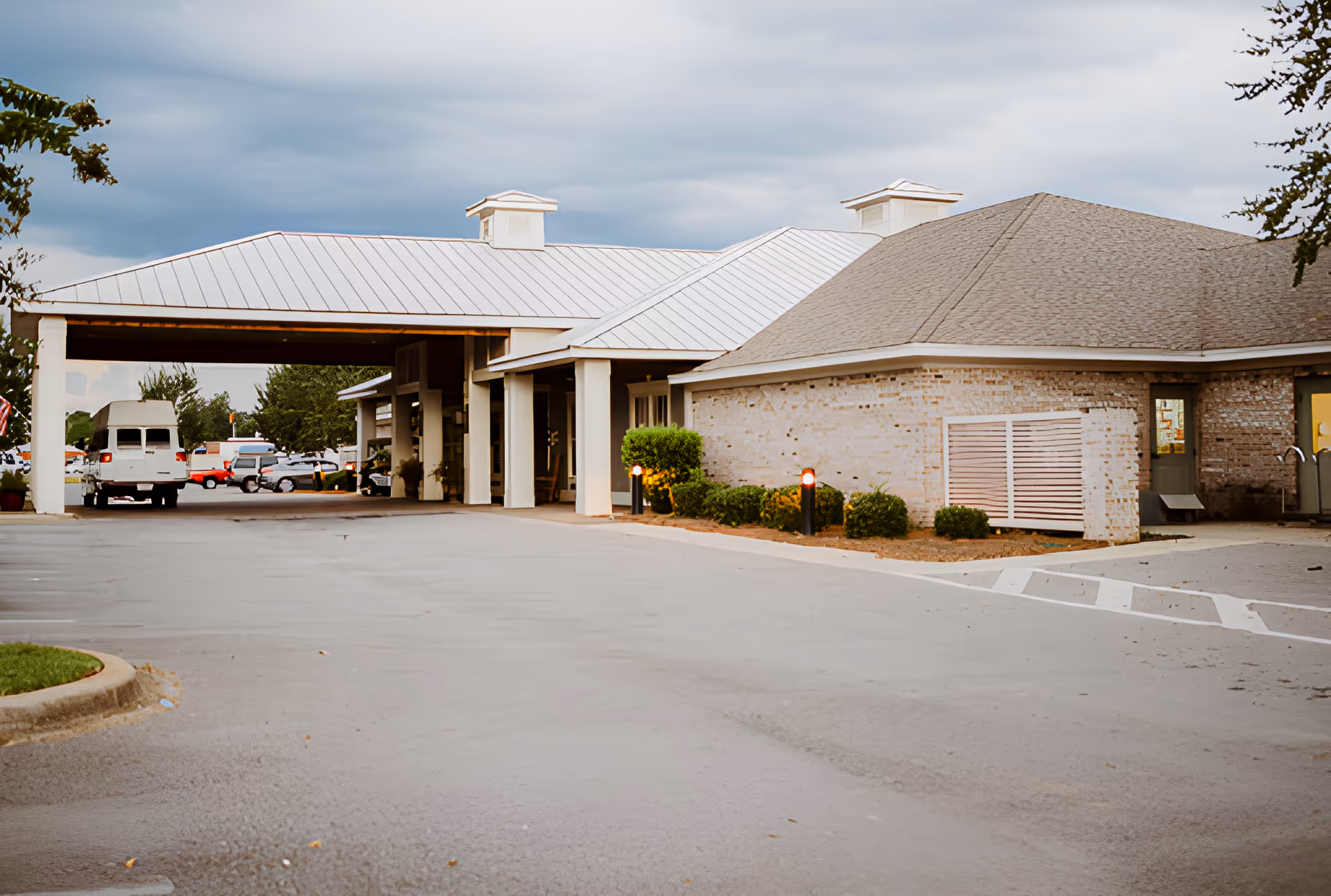 Exterior view of The Terrace At Ivey Acres Assisted facility showing a covered entrance with a metal roof, a brick building with a shingled roof, some bushes, and a parking area with several vehicles.