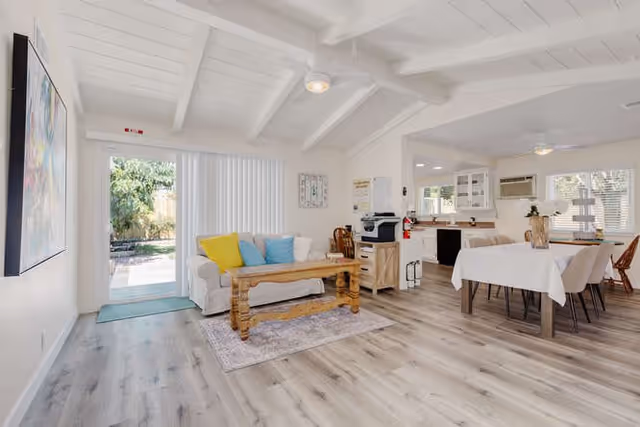 A bright and spacious interior room featuring a white couch with colorful cushions, a wooden coffee table on a small rug, a dining table with white tablecloth and chairs, and a kitchen area with white cabinets and appliances. Large windows and a glass door allow natural light to fill the space, showing a glimpse of greenery outside.