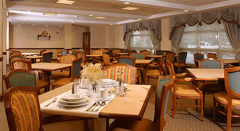 A spacious dining room with multiple wooden tables and chairs arranged neatly. Each table is set with plates, cups, silverware, and a centerpiece of dried flowers. Large windows with decorative curtains allow natural light to fill the room.